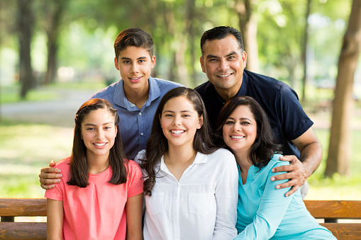 A latin family of five sitting and standing together, embracing and smiling at the camera in a horizontal waist up shot outdoors.